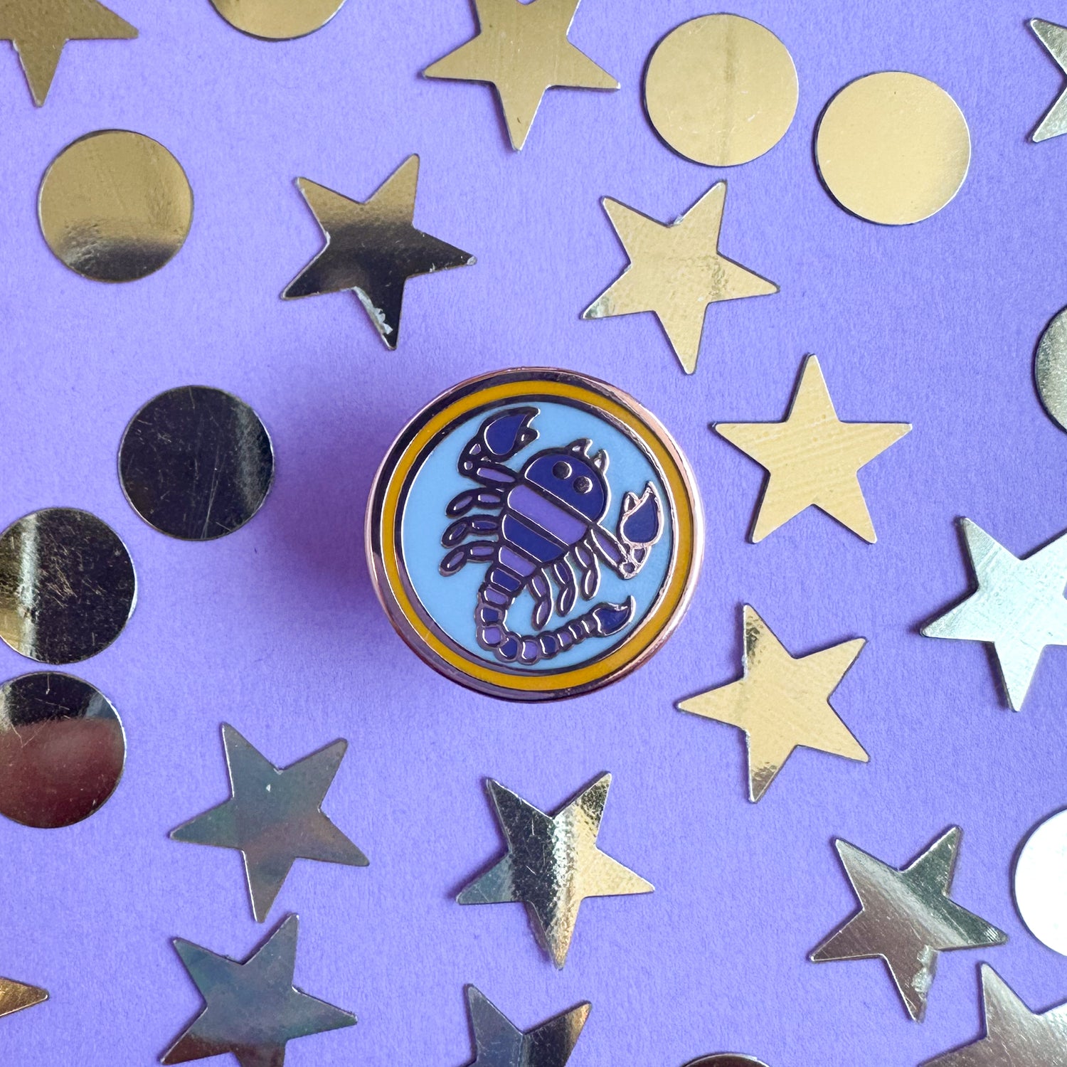 A circular enamel pin with a purple scorpion on it to represent the Scorpio zodiac sign. The pin is on a purple background with gold stars and circles around the pin. 
