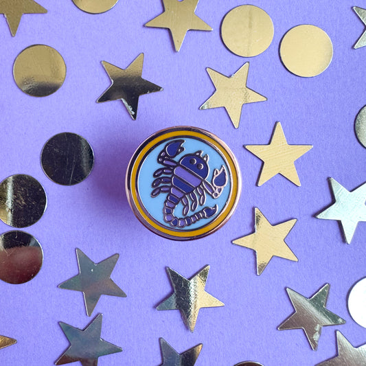 A circular enamel pin with a purple scorpion on it to represent the Scorpio zodiac sign. The pin is on a purple background with gold stars and circles around the pin.