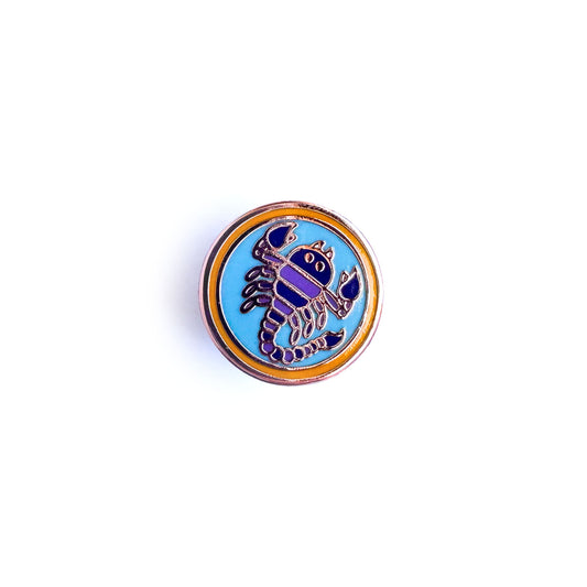 A circle enamel pin with a scorpion in a blue circle inside an orange circle meant to symbolize the Scorpio zodiac sign.
