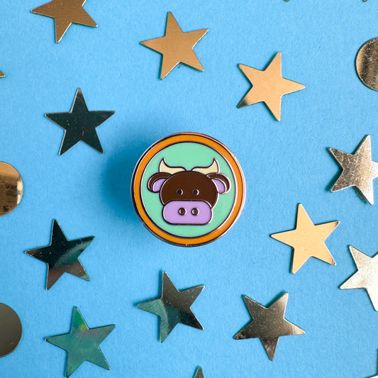 A pin of a circle with a cute bull illustration in it meant to represent the Taurus zodiac sign. The pin is on a blue background surrounded by gold star and circle confetti. 