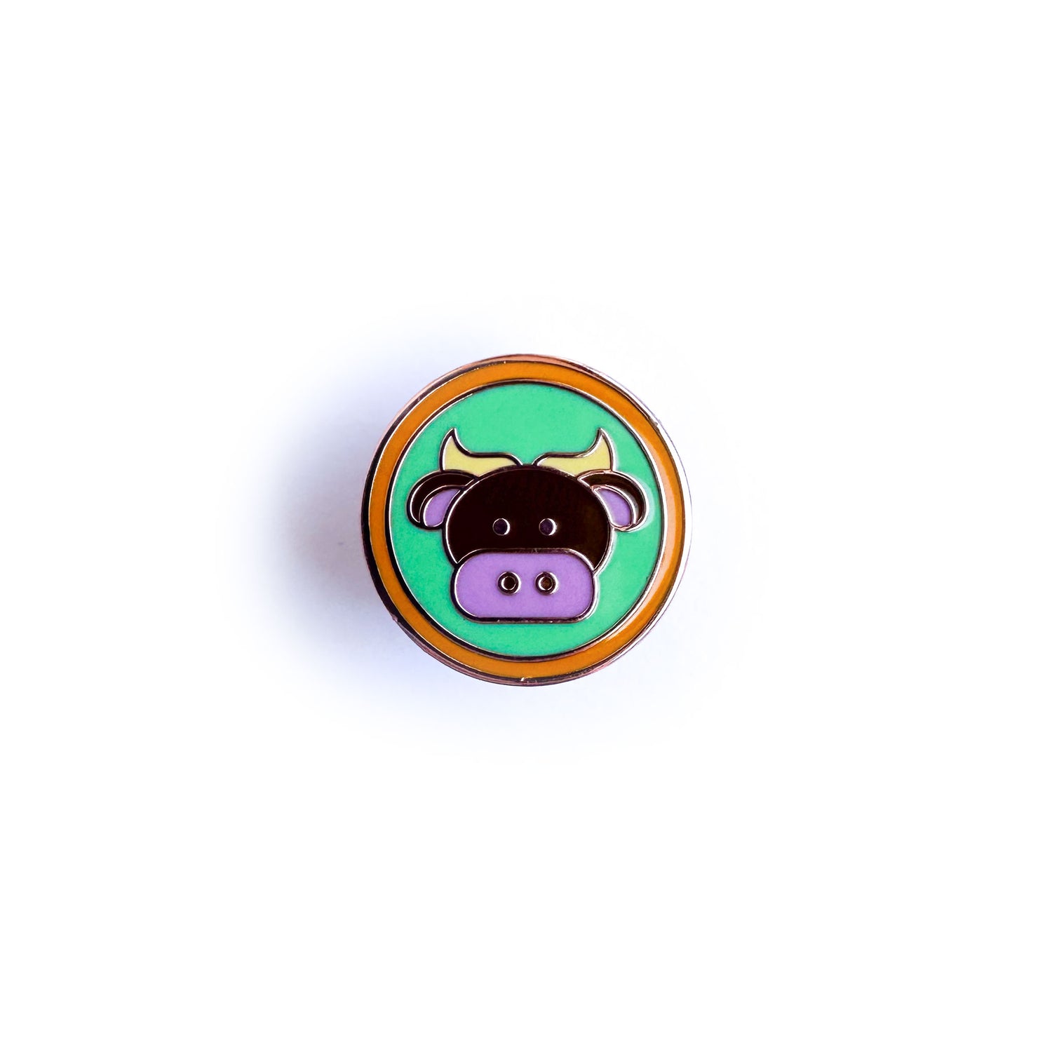 An enamel pin of a green circle with an orange border with a cute brown bull inside the green circle. The bull is meant to represent the Taurus Zodiac sign.