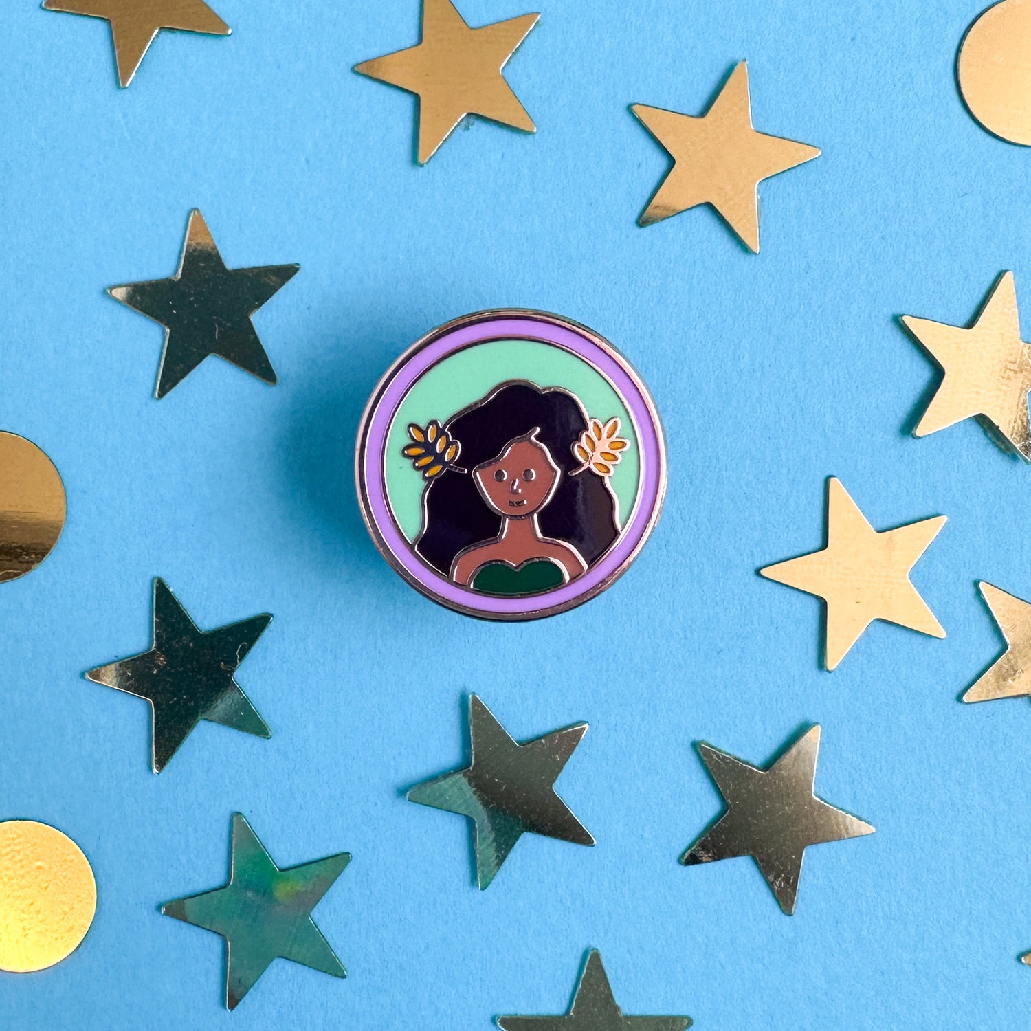 A circle enamel pin with an illustration of a woman with brown skin and flowing brown hair with wheat fronds in it to represent the Virgo zodiac sign symbol. The pin is on a blue background with gold star and circle confetti surrounding it. 