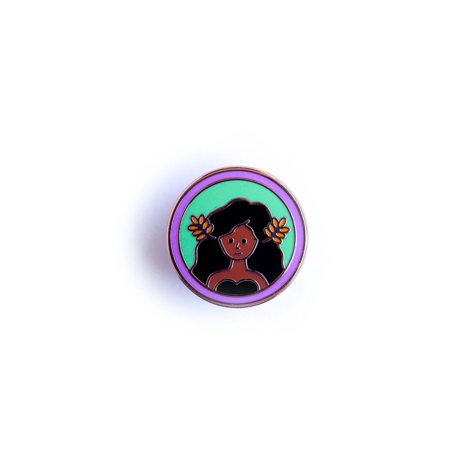 An enamel pin of a woman with brown skin with flowing brown hair with wheat fronds in it. She is in a green circle surrounded by a lavender circle. 