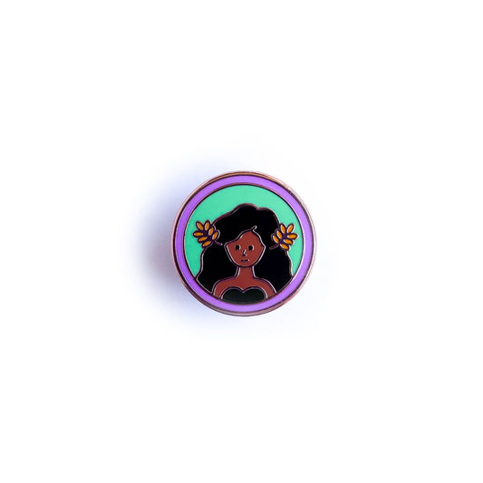 An enamel pin of a woman with brown skin with flowing brown hair with wheat fronds in it. She is in a green circle surrounded by a lavender circle.