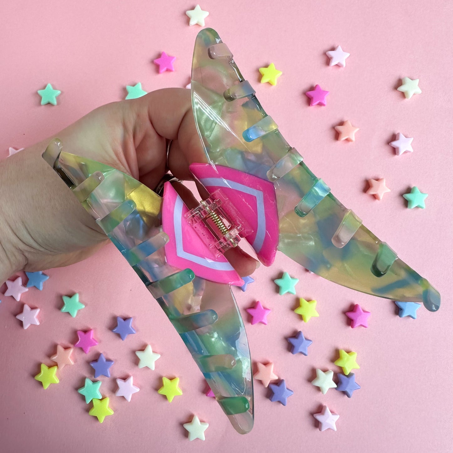 A hand opening a large claw clip made out of pastel acetate. The background of the image is pink with plastic star beads around it.