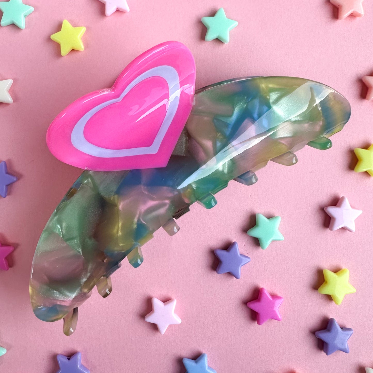 A claw clip with swirled pearly acetate in pink, blue, and yellow with a heart shaped handle. The clip is on a pink background with pastel star beads scattered around it. 