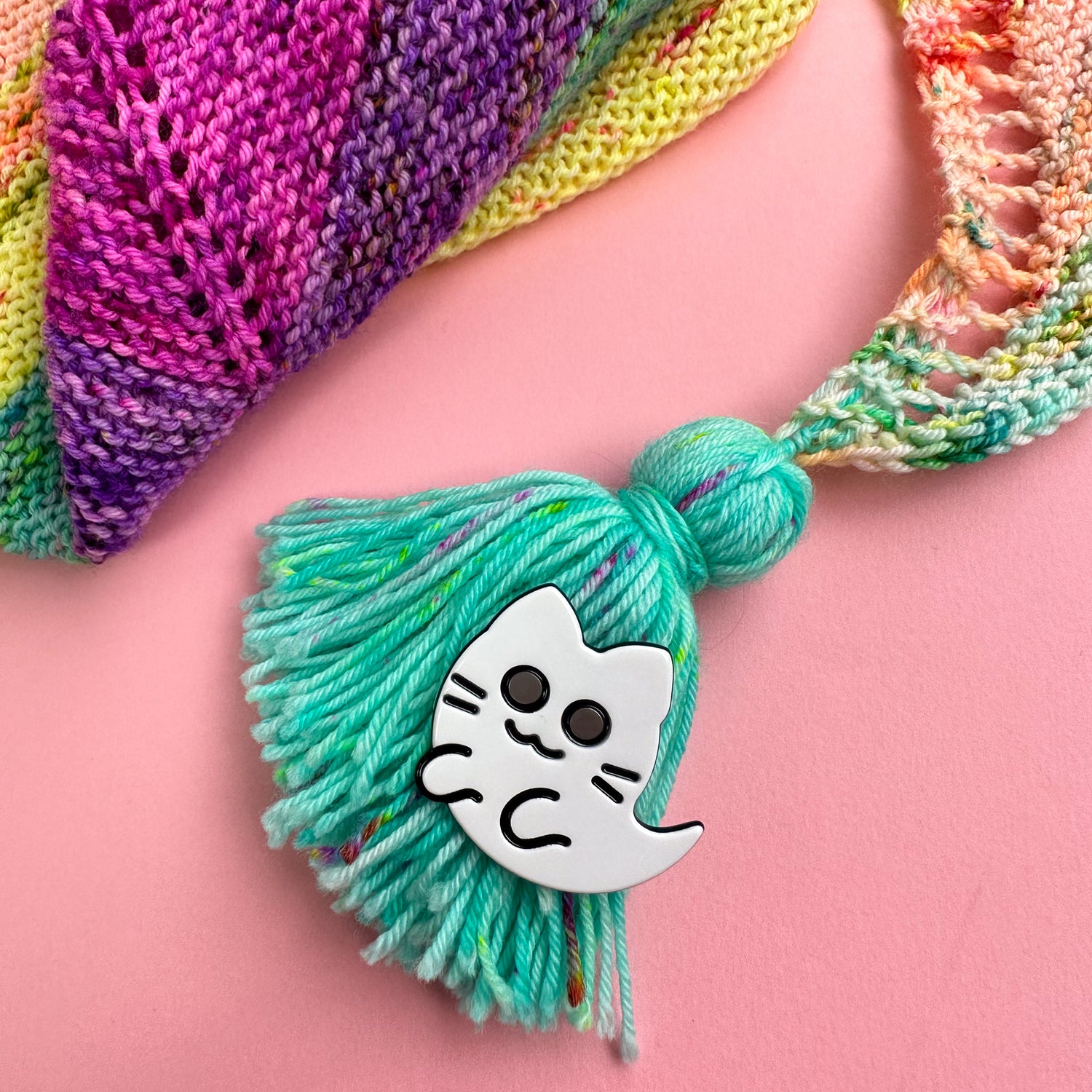 A ghost kitty hair clip attached to a turquoise handmade tassel that is attached to a rainbow hand knit shawl in the corner of the image. 