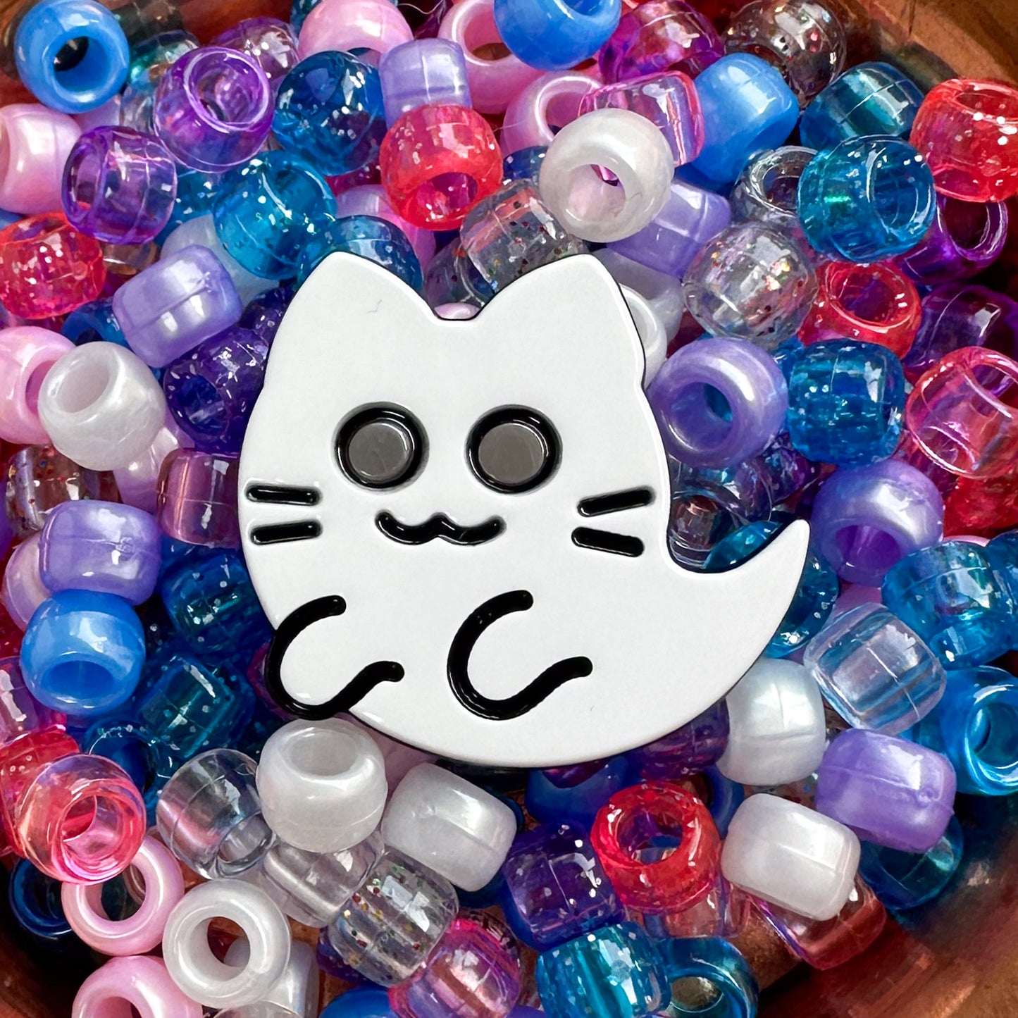 A close up of the Ghost Kitty Babette showing it sitting in a pile of blue, pink, and purple pony beads. 