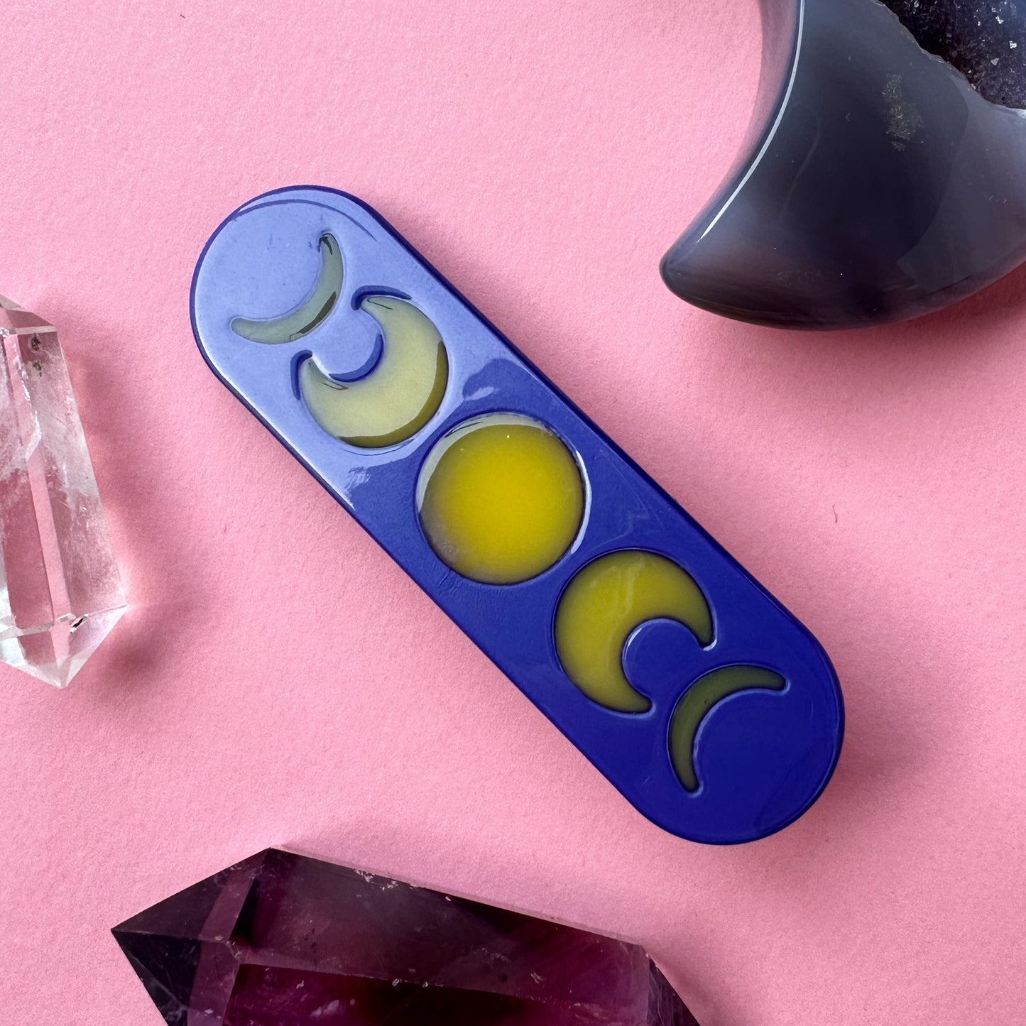 An acetate hair clip that is purple with yellow moon phases on it on a pink background surrounded by crystals.