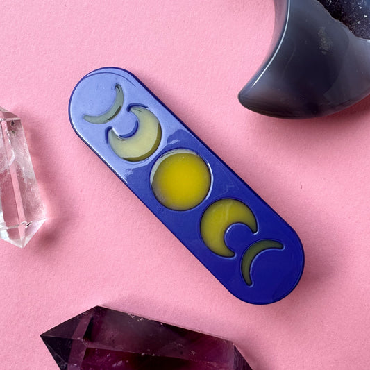 An acetate hair clip that is purple with yellow moon phases on it on a pink background surrounded by crystals.