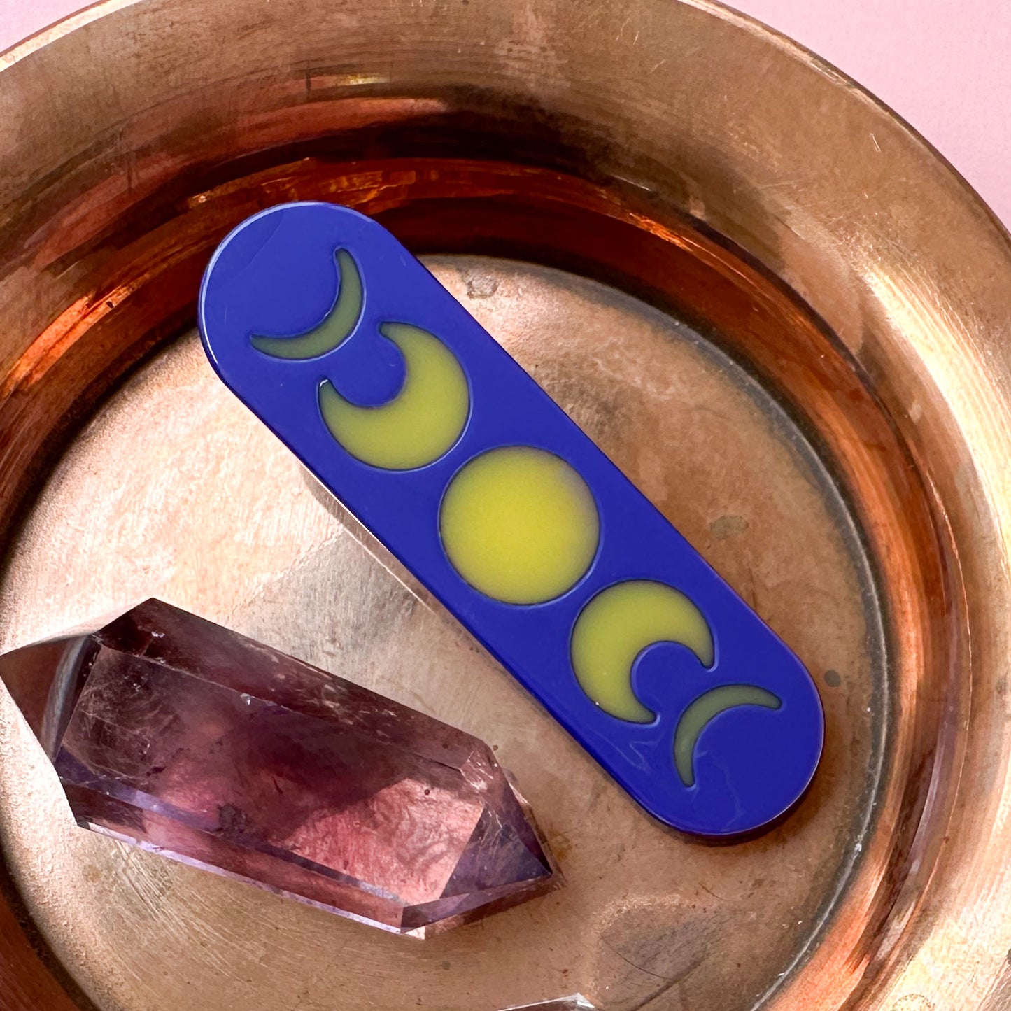 A hair clip that is shaped like a bandaid and is a purple background with shapes of moon phases on it. There is a double pointed amethyst crystal next to it and both items are in a copper dish. 