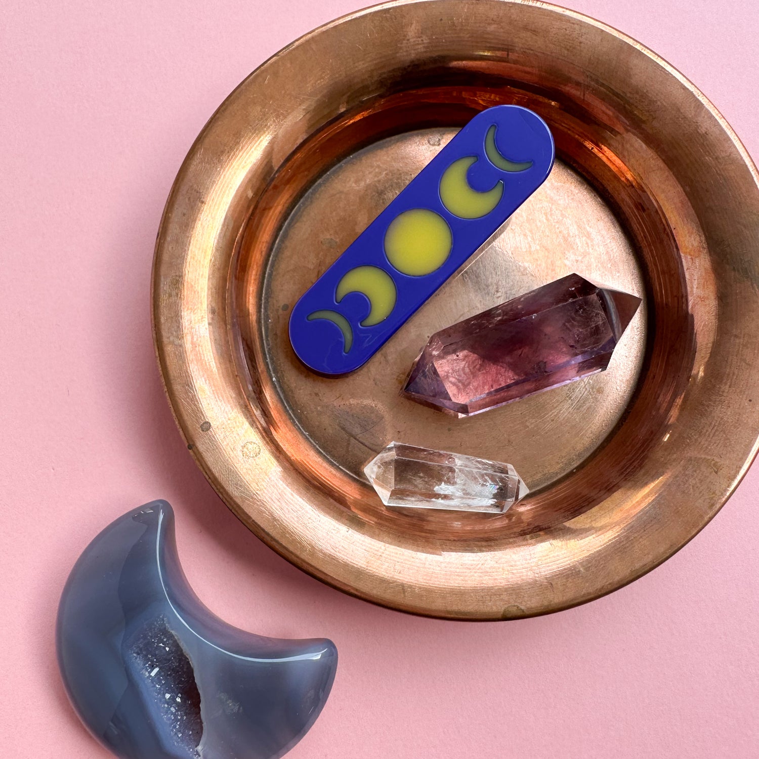 A purple hair clip with yellow moon phases on it in a copper bowl surrounded by double point crystals and a moon shaped crystal. 