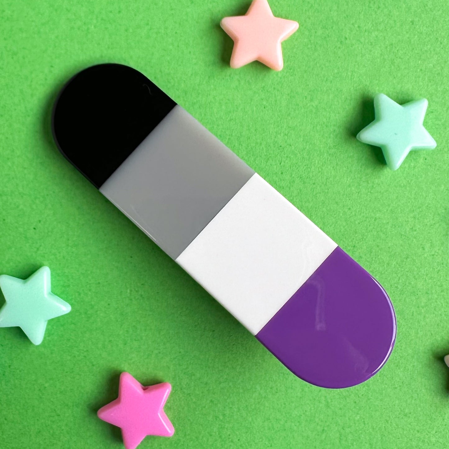 A capsule shaped acetate hair clip with vertical stripes of the Asexual Pride Flag. The clip is on a green background with plastic star beads around it. 