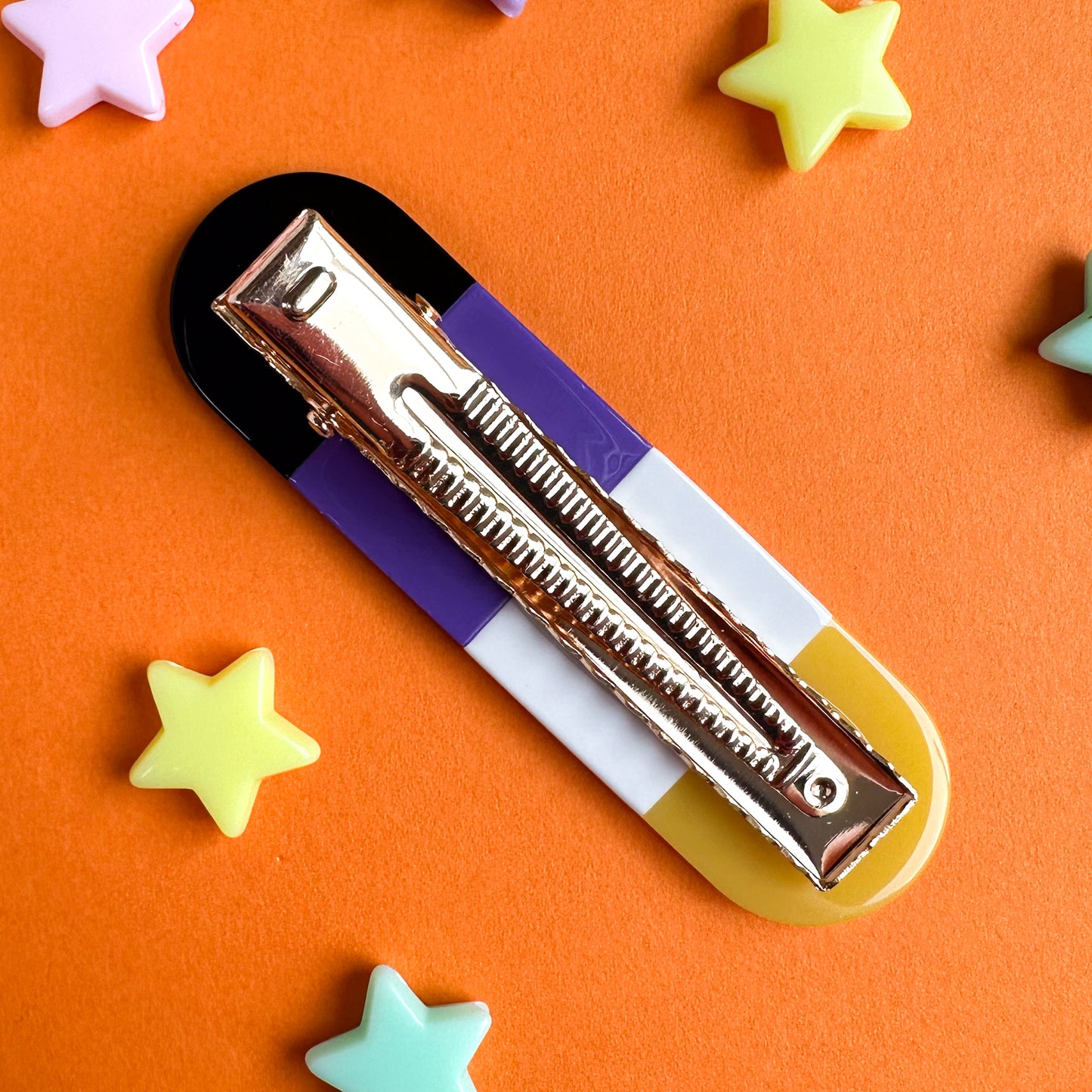 An alligator style hair clip with a gold clip mechanism. The body o the clip is the colors of the Nonbinary Pride Flag. The clip is on an orange paper background with pastel star beads scattered around it. 