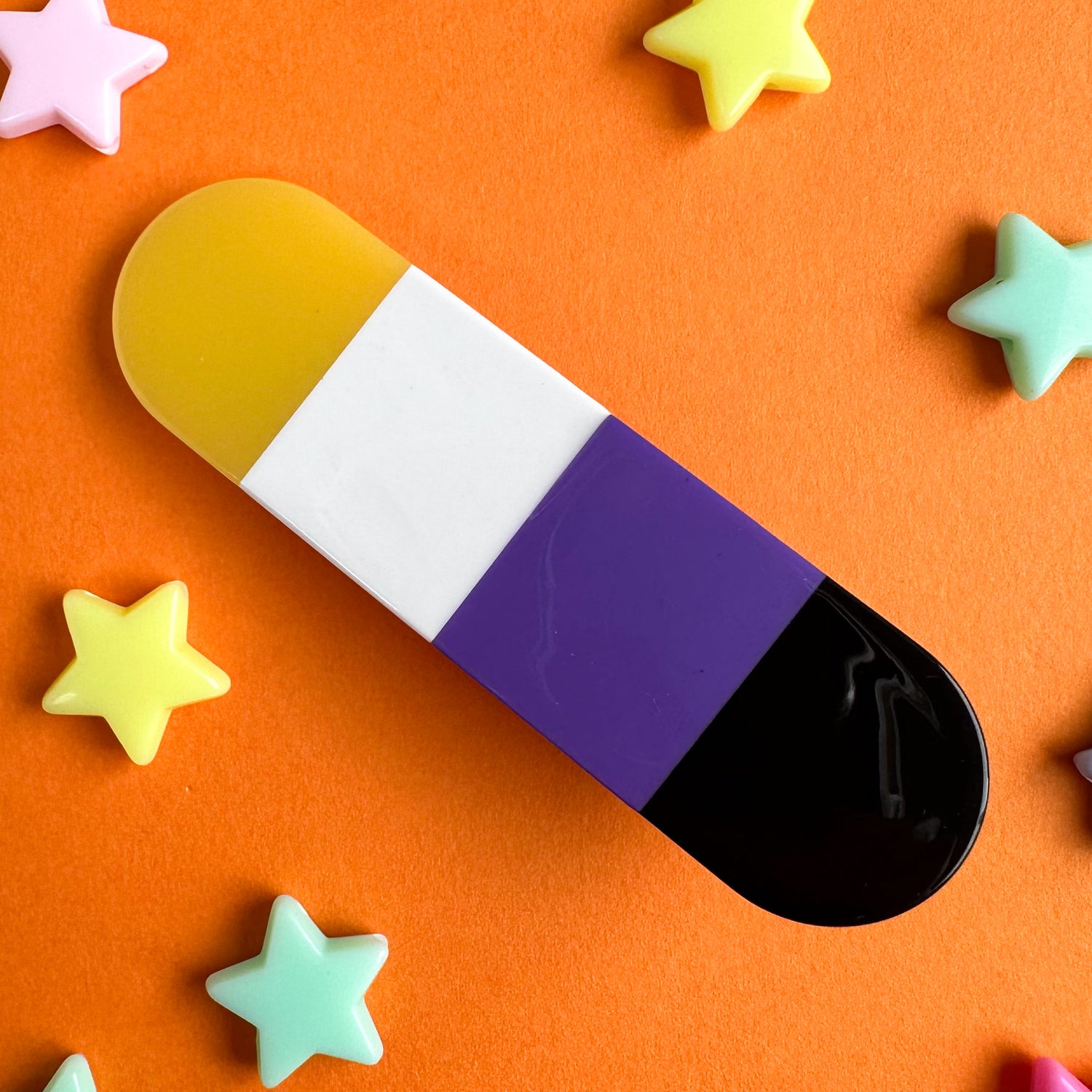 A capsule shaped hair clip in the colors of the Nonbinary Pride Flag. The hairclip is on an orange background with plastic star beads around it. 