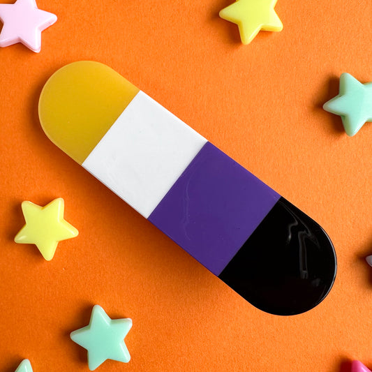 A capsule shaped hair clip in the colors of the Nonbinary Pride Flag. The hairclip is on an orange background with plastic star beads around it. 