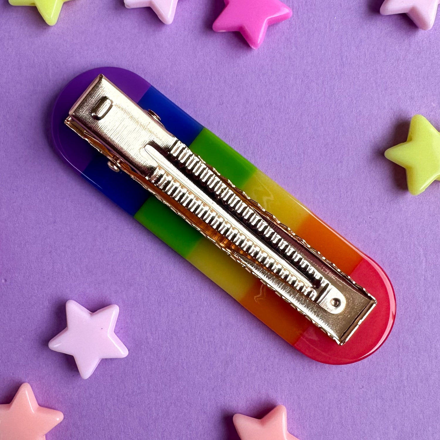 A rainbow hair clip with a gold alligator style clip. The clip is on a purple background with star beads around it. 