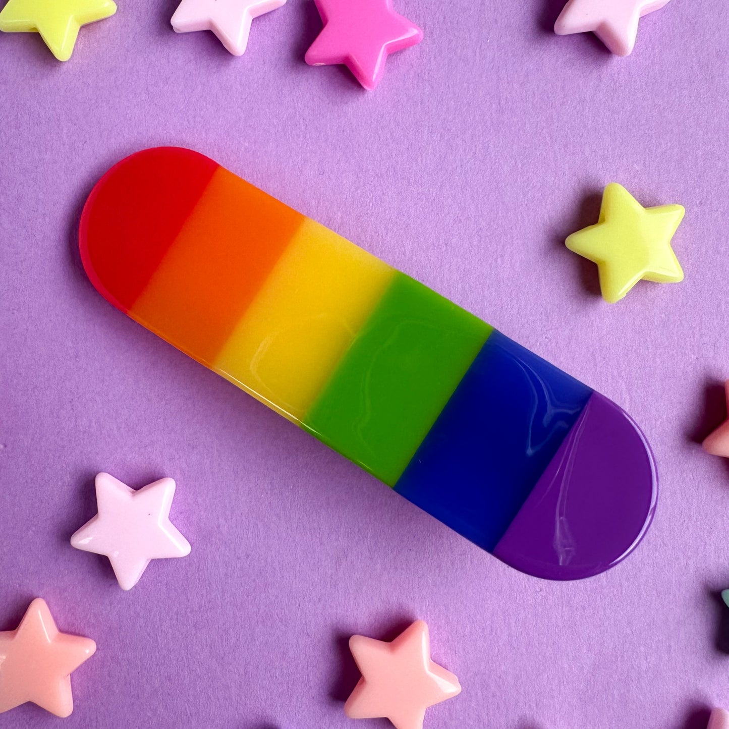 A capsule shaped hair clip with vertical stripes in the colors of the rainbow. The hair clip is on a purple background with pastel star beads around it. 