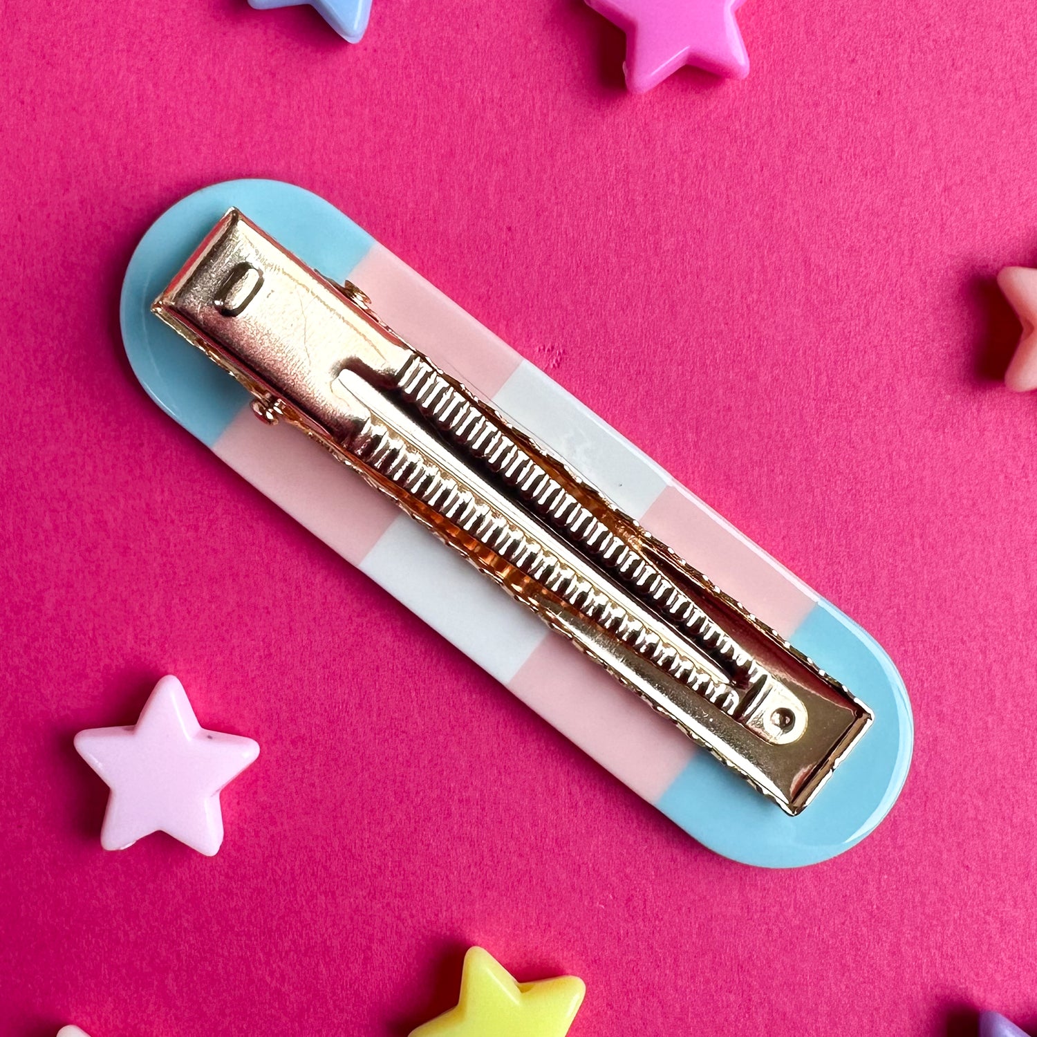 A gold alligator style clip with a base in the colors of the transgender pride flag. The clip is on a pink background with star beads around it. 