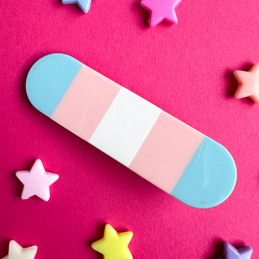 A capsule shaped hair clip with vertical stripes in the colors of the transgender pride flag. The clip is on a hot pink paper background with pastel stars around it.