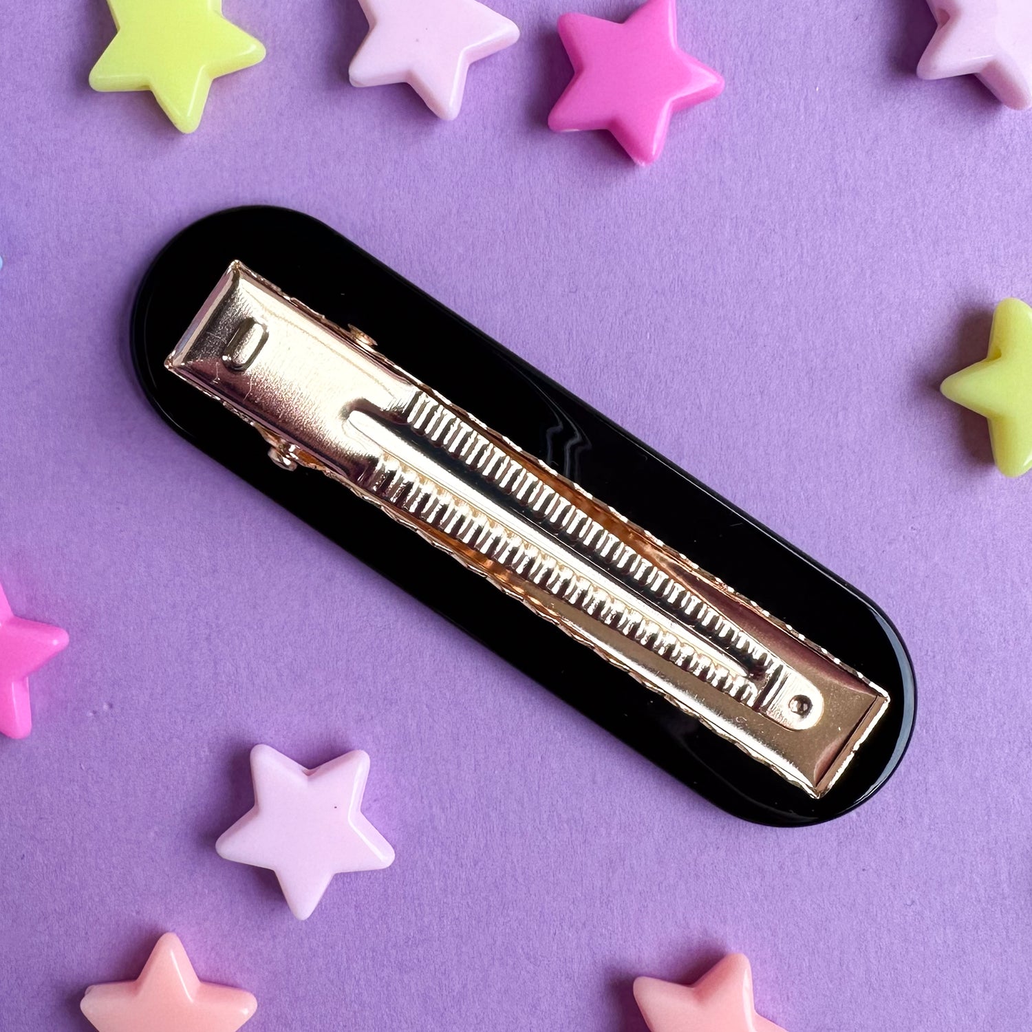 A black oval shaped hairclip with a gold alligator shaped clip. A purple paper background with pastel star beads around it. 