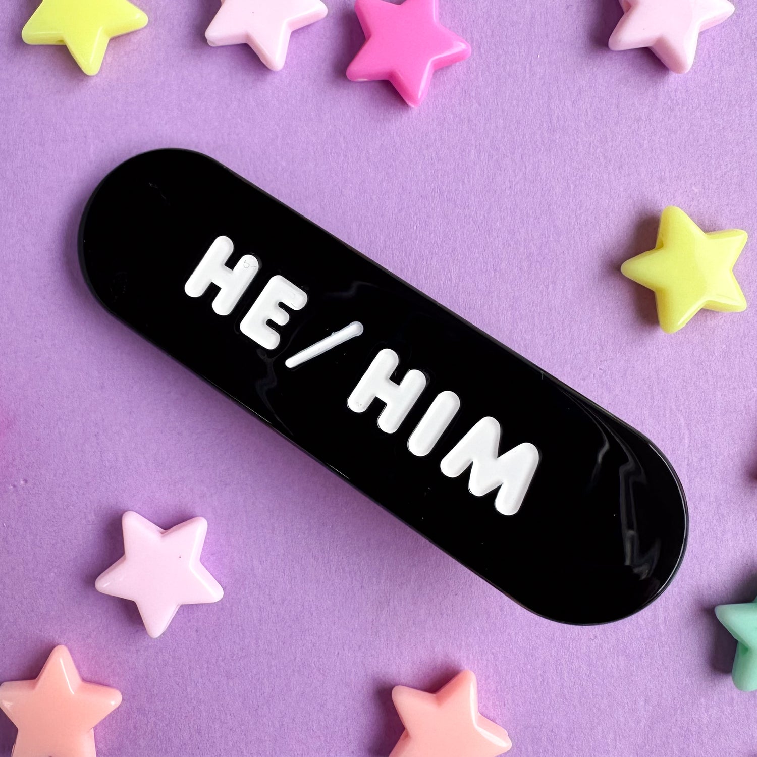 A black capsule shaped hairclip with embossed words on it that read "He/Him". The pin is on a purple piece of paper with pastel star beads around it. 