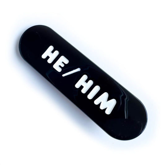 A black capsule shaped hairclip with embossed words on it that read "He/Him"