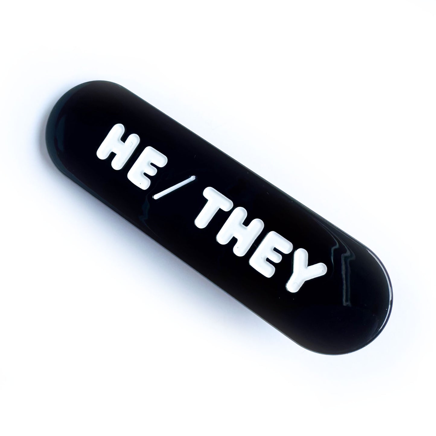 A black capsule shaped hairclip with embossed words that read "He/They" on it. 