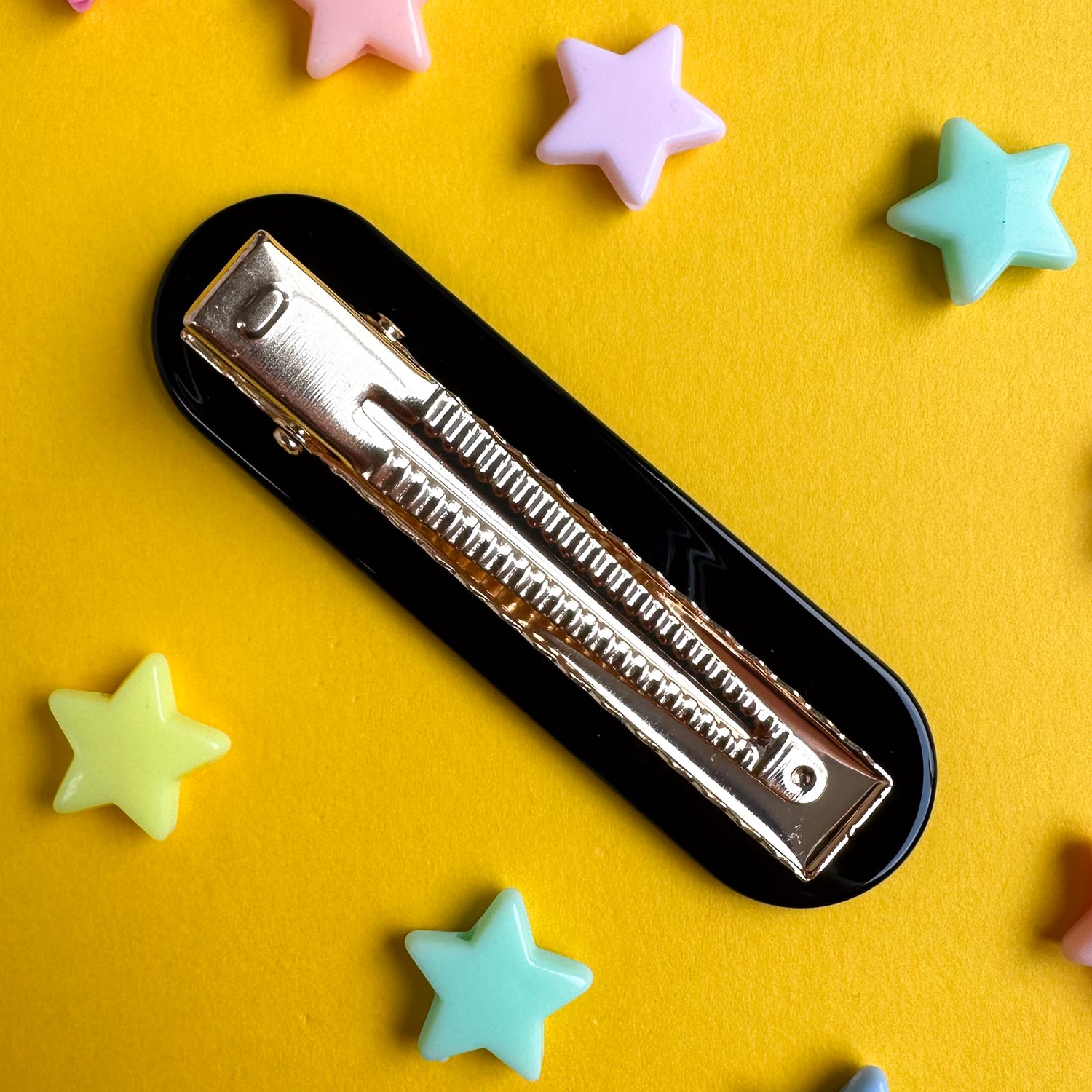 An alligator hair clip with a black clip part. The hair clip is on a yellow paper background with plastic beads shaped like stars around it. 