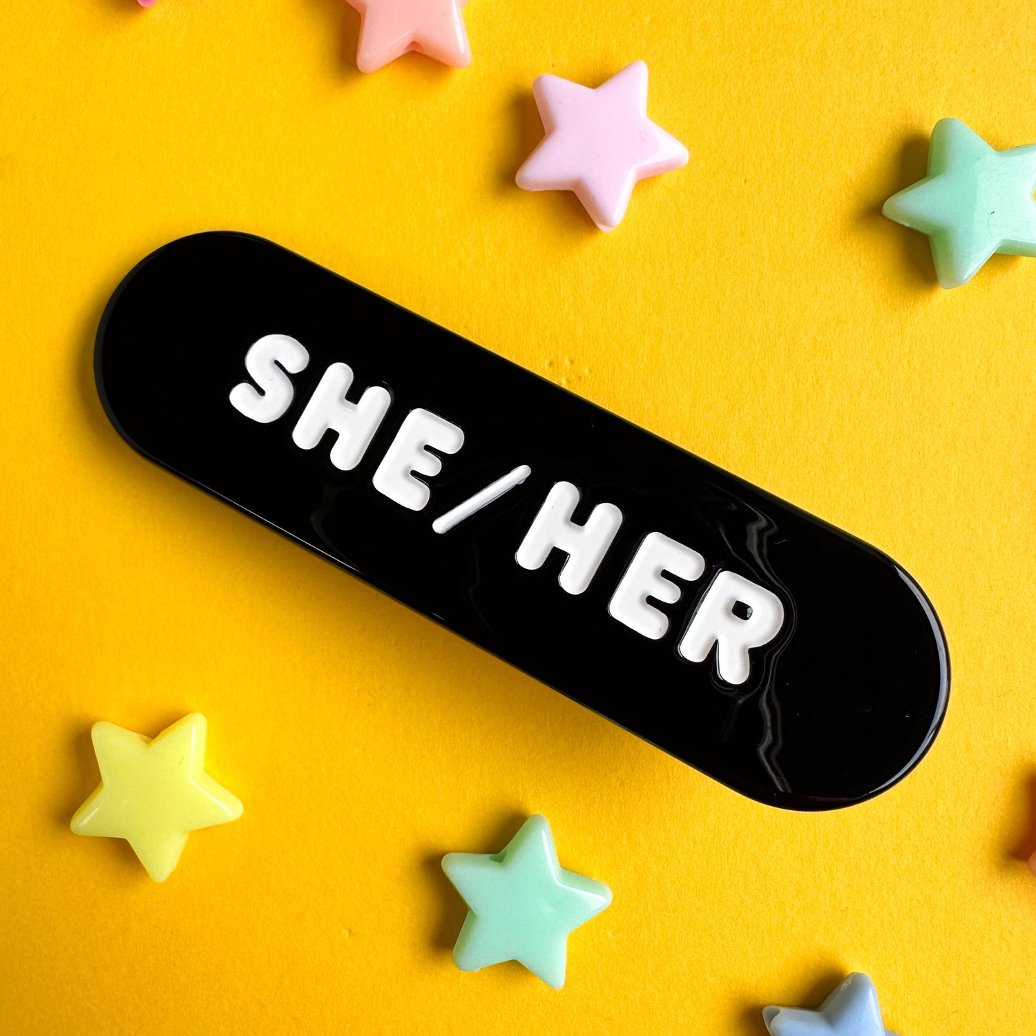 A black capsule shaped hair clip with embossed white words that read "She/Her".  The hair clip is on a yellow paper background with plastic star beads around it. 