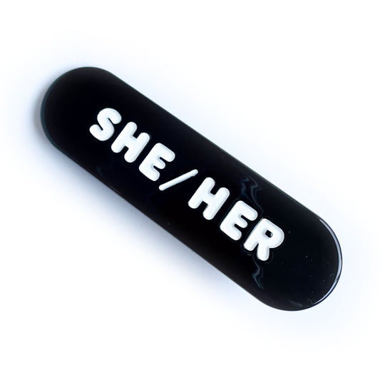 A black capsule shaped hair clip with embossed white words that read "She/Her".