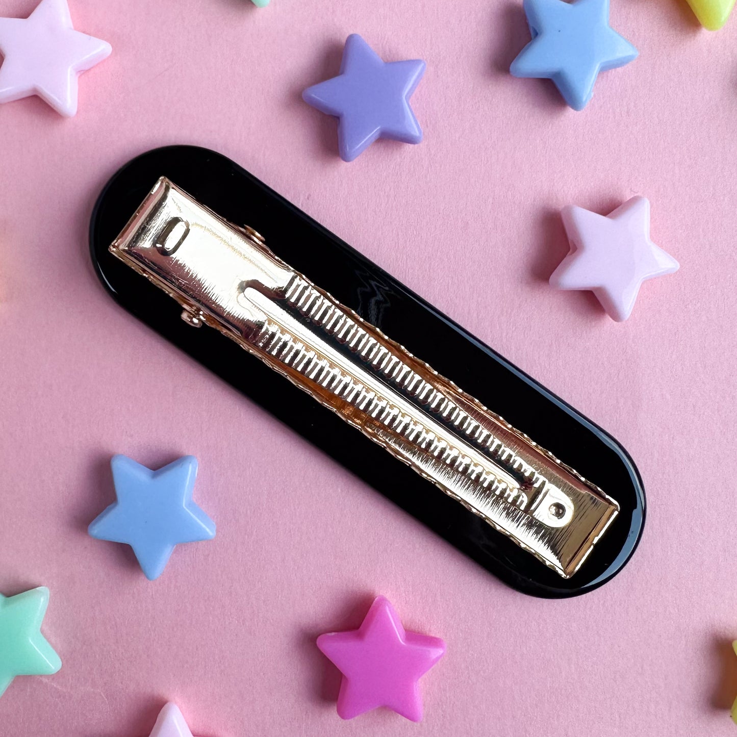 A gold alligator style hair clip with a black base. It's sitting on a pink paper background with plastic star beads around it. 