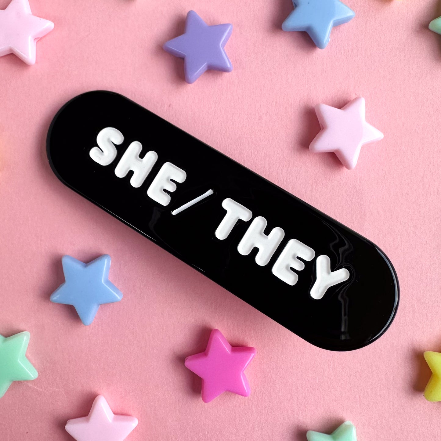 A black capsule shaped hair clip with embossed white words that read "She/They". The clip is on a pink background with plastic star beads around it. 