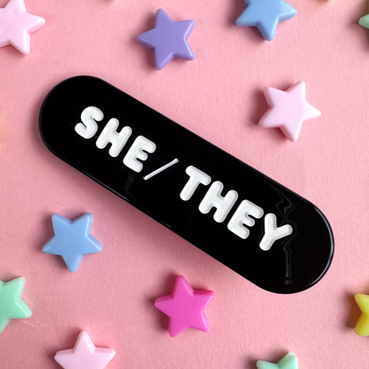 A black capsule shaped hair clip with embossed white words that read "She/They". The clip is on a pink background with plastic star beads around it.