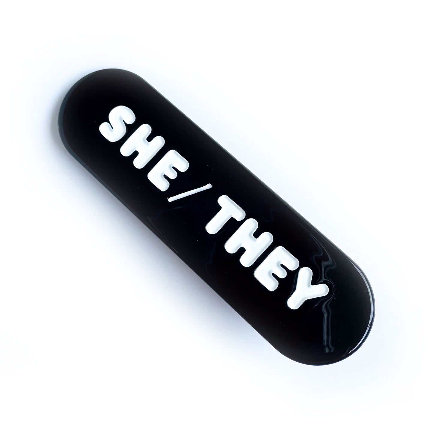 A black capsule shaped hair clip with embossed white words that read "She/They".