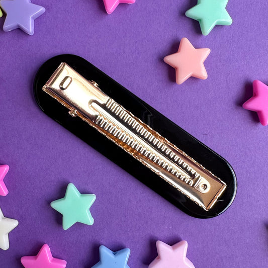 An alligator style gold clip with a black base. The clip is on a purple paper background with pastel star beads scattered around it. 