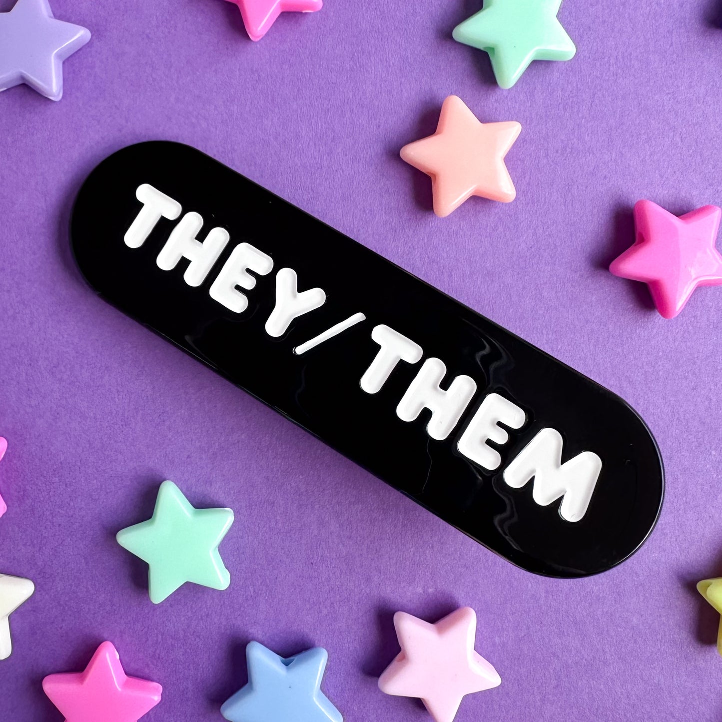 A black capsule shaped hair clip with embossed white letters that read "They/Them".  The clip is on a purple background with plastic star beads around it. 