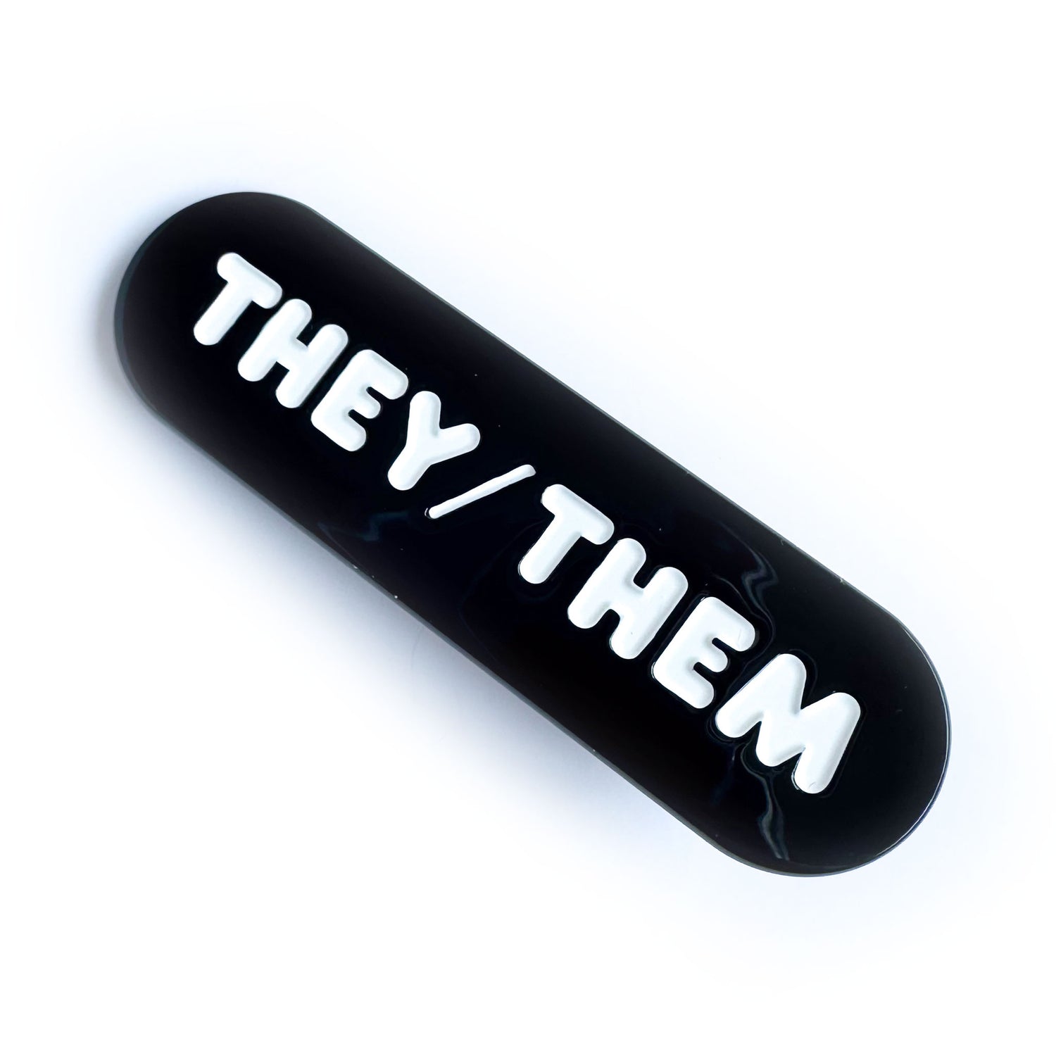 A black capsule shaped hair clip with embossed white letters that read "They/Them". 