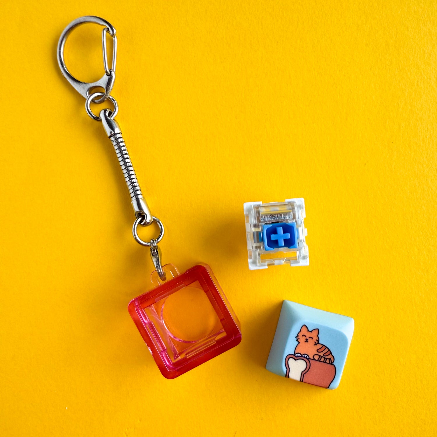 A clicky cat keychain with the Gateron Blue Switch removed from the clear hot pink housing. The blue keycap with an orange cat in loaf position on top of a loaf of bread is next to these things. 
