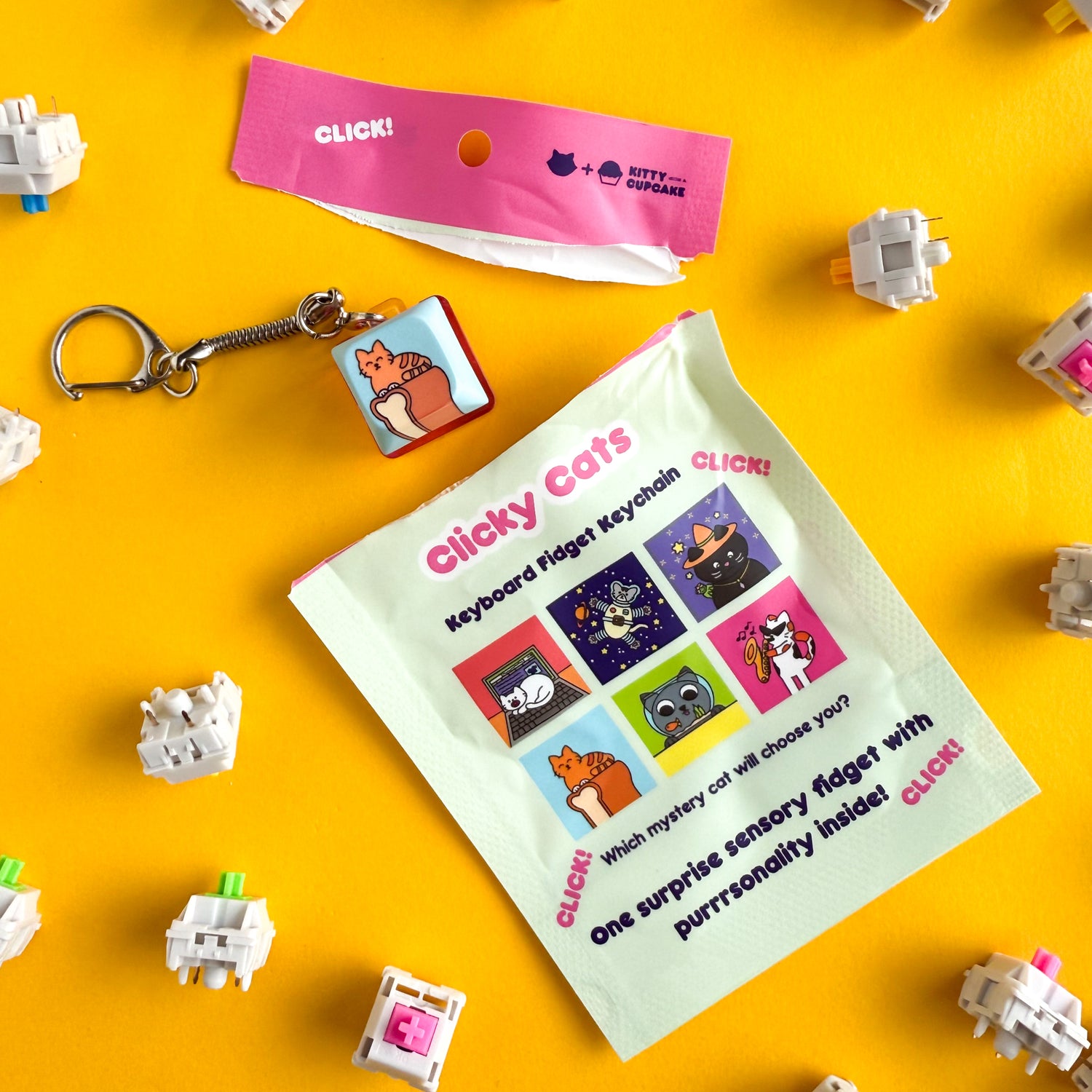 A torn open blind bag revealing a keychain clicker inside with an illustration of an orange cat in loaf position on top of a loaf of bread. The blind bag reads "Clicky Cats Keyboard Fidget Keychain". There are keyboard switches scattered around on top of a yellow background. 