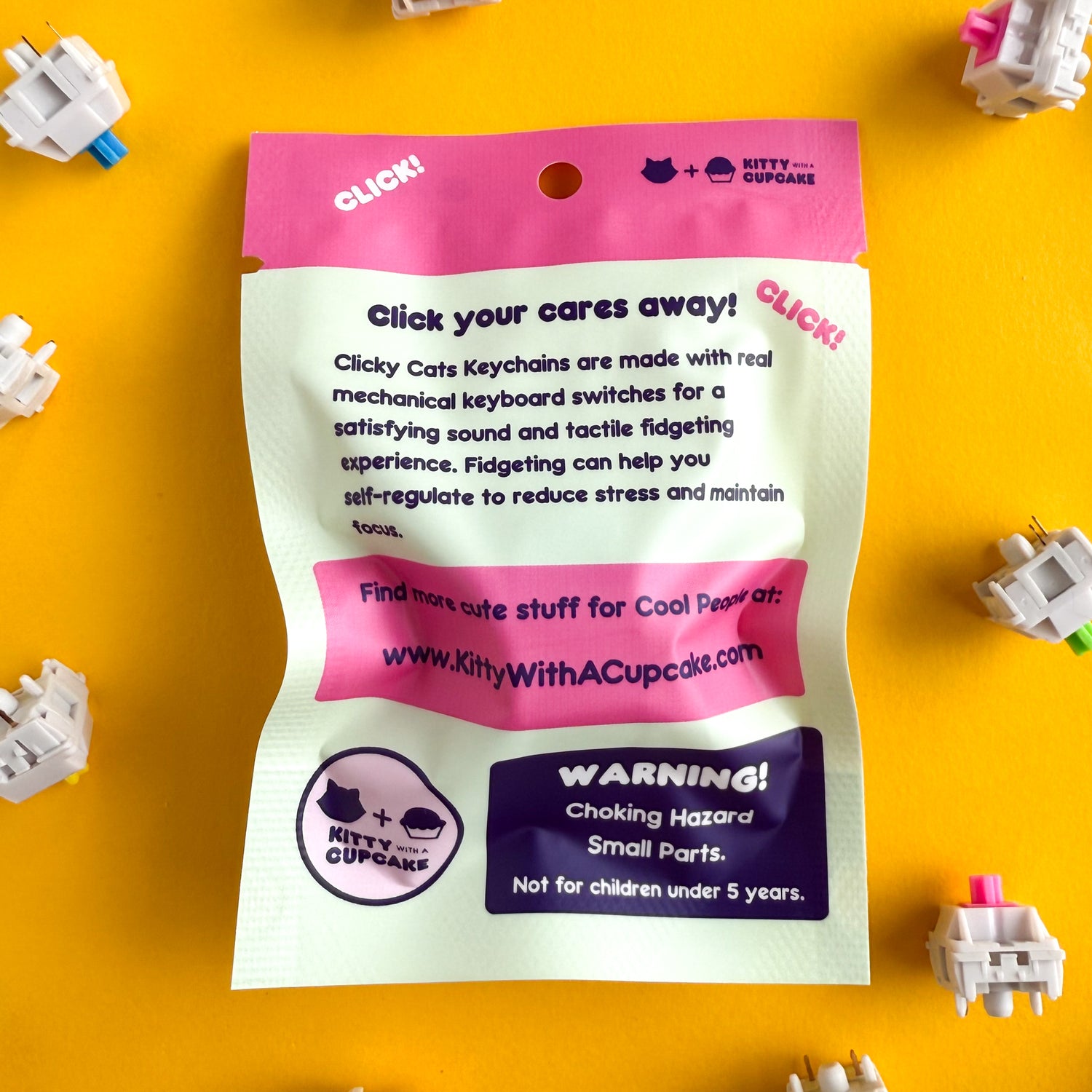 The back of the Clicky Cats packaging. It reads "Click your cares away! Clicky Cats Keychains are made with real mechanical keyboard switches for a satisfying sound and tactile fidgeting experience. Fidgeting can help you self regulate to reduce stress and maintain focus. Find more cute stuff for Cool People at: www.kittywithacupcake.com WARNING! Choking Hazard. Small Parts. Not for children under 5 years." The bag is mint with hot pink details. It is on a yellow background with keyboard switches around. 