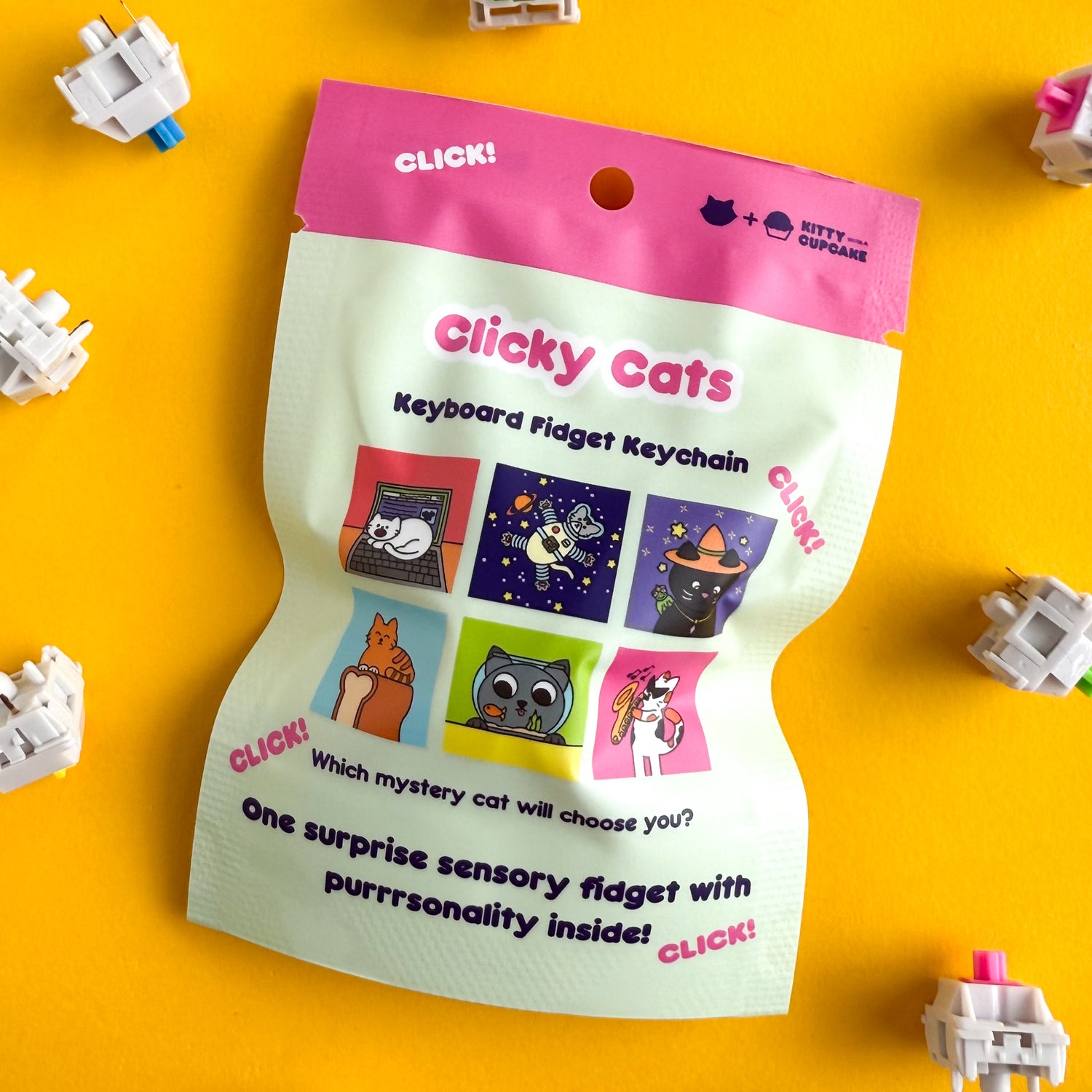 A mint colored bag with a hot pink top that reads "Clicky Cats - Keyboard Fidget Keychain" There are cartoon illustrations of a different cats on the package. The cats are a white cat on a laptop, an astronaut cat, a witch cat, a loaf cat, a fishbowl cat, and a jazz cat. There are keyboard switches around the bag. Everything is on a yellow background. 