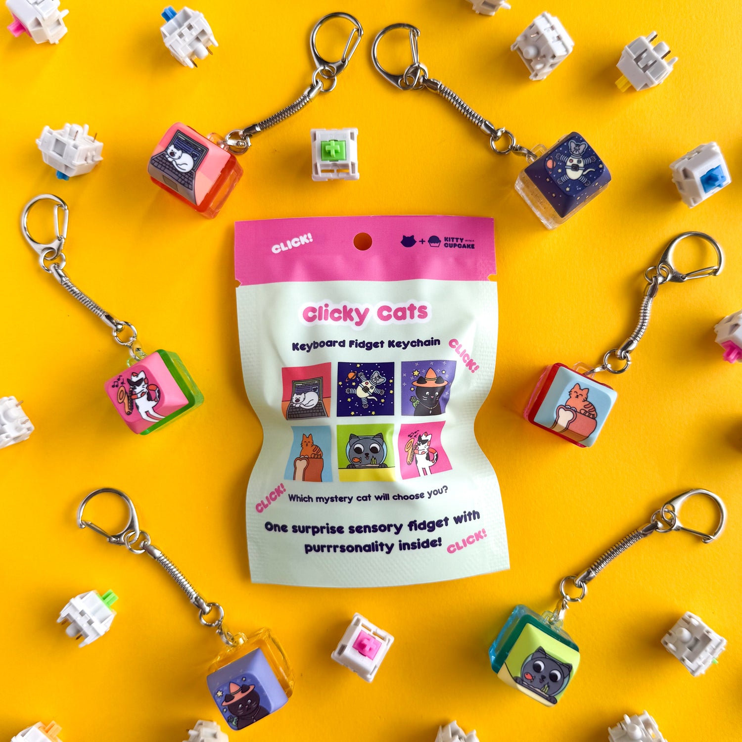 An opaque mint bag with a hot pink top reads "Clicky Cats - Keyboard Fidget Keychain" and has pictures of square illustrations of cats below. There is a corresponding keycap clicky keychain to each of the illustrations positioned around the bag. There is an astronaut cat in space, an orange cat on a loaf of bread, a grey cat looking at a goldfish in a bowl, a black cat in a witch hat, a calico cat in sunglasses with a saxophone, and a white cat on a laptop. There are mechanical keyboard switches scattered. 