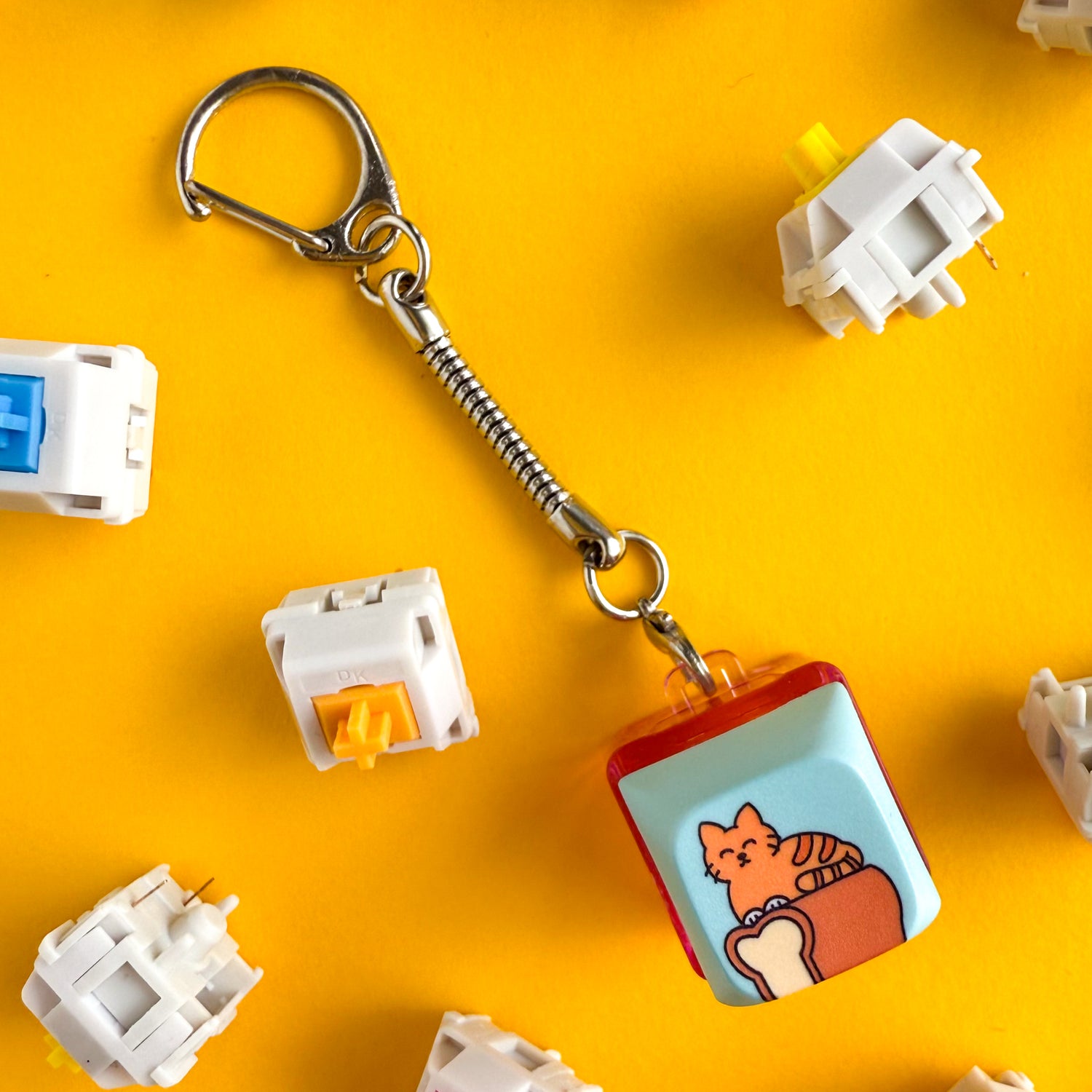 A keychain attached to a keycap fidget clicky toy. The keycap has a cartoon illustration of an orange cat in loaf position on top of a loaf of bread, the background is light blue. The base of the keyboard keychain is hot pink. There are mechanical keycap switches around the keychain. Everything is on a yellow background. 