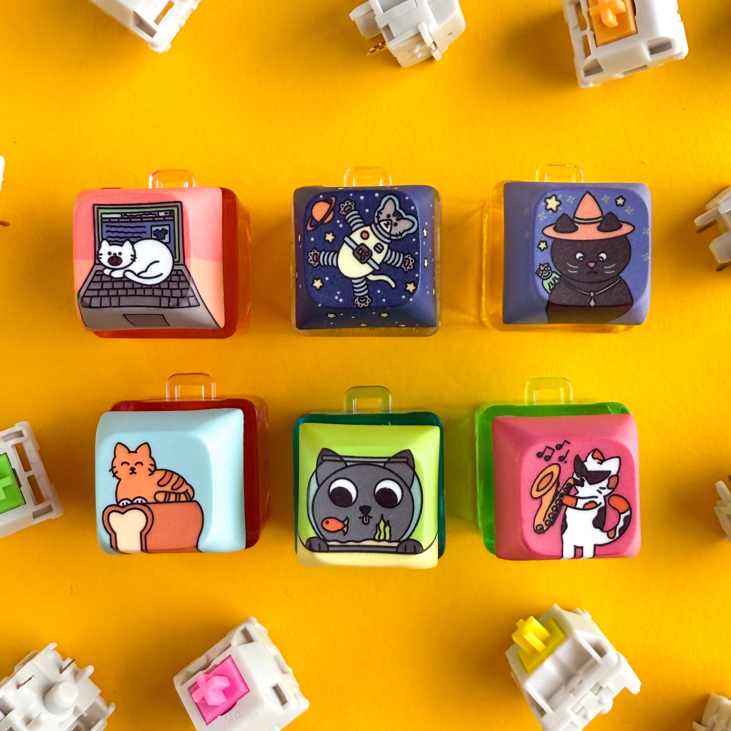 Six cube shaped keycap clicky keychains. All of them have bright colors and feature cute illustrations of cats. One of them is a white cat on a laptop, one is a grey cat in an astronaught suit in space, one is a black cat with a witch hat and a frog on it's  shoulder, one is an orange cat on a loaf of bread, one is a grey cat looking at a gold fish in a bowl, and one is a calico cat with sunglasses playing a saxophone. The items are on a yellow background with mechanical keyboard switches around. 