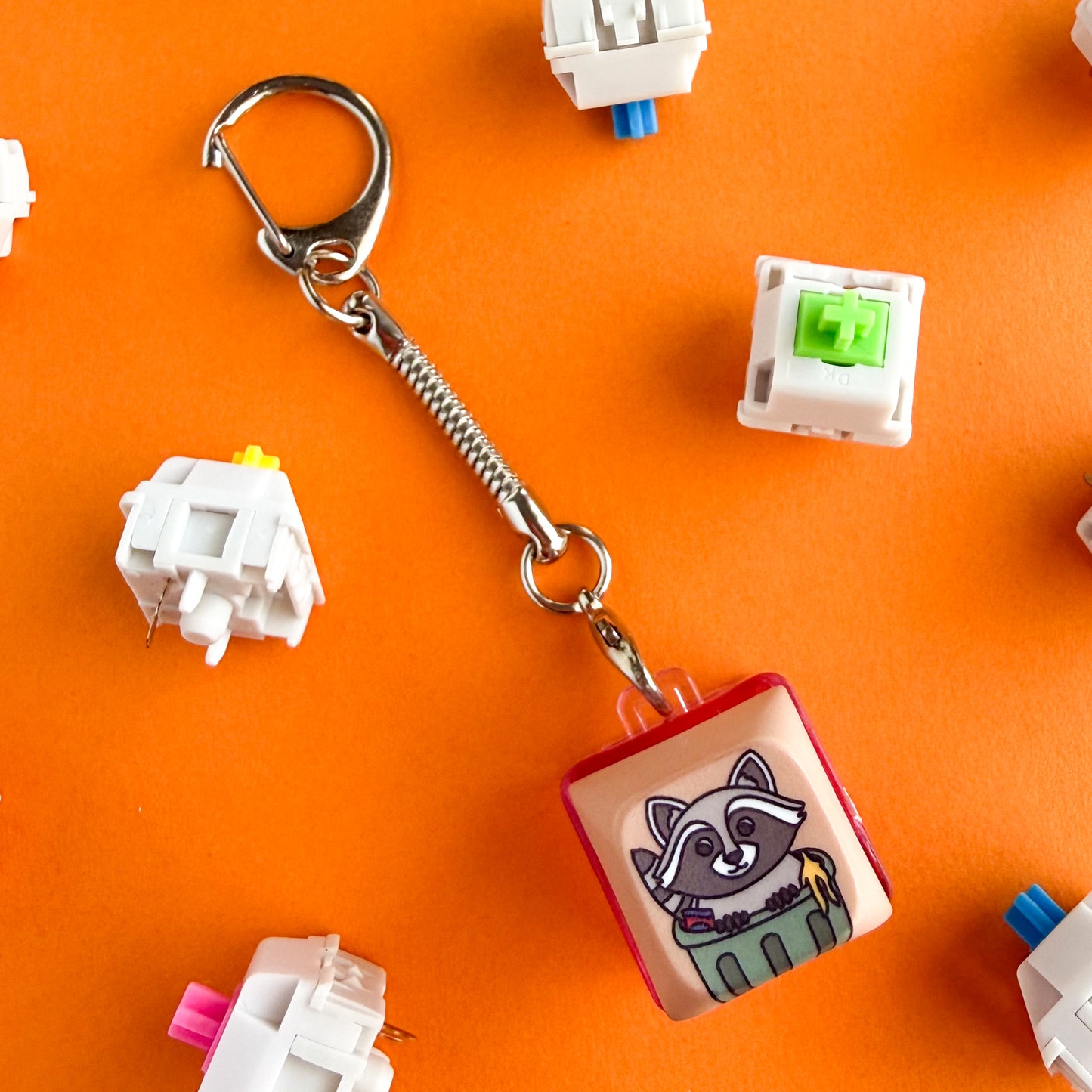 A fidget clicker keychain with a cute cartoon of a raccoon in a trash can with a banana peel on the keycap. The base of the keychain is hot pink. The keychain is on an orange background and there are color keyboard switches around it. 