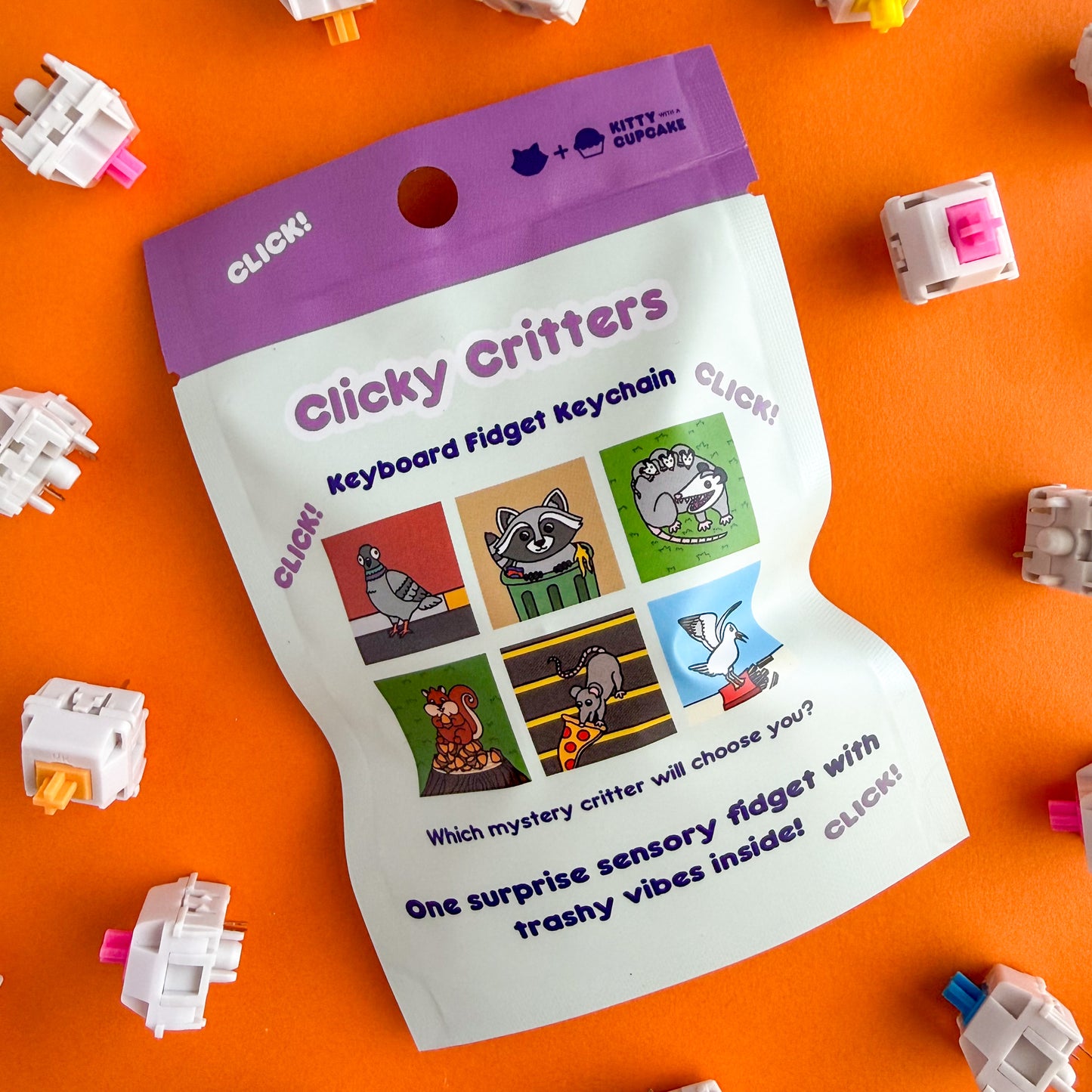 A blind bag that is mostly mint with a purple top. It reads "Clicky Critters Keyboard Fidget Keychain" and has pictures of what designs you could get in the bag including a pidgeon, raccoon, opossum, squirrel, pizza rat, and seagull. The bag is on an orange background with keyboard switches around it. 