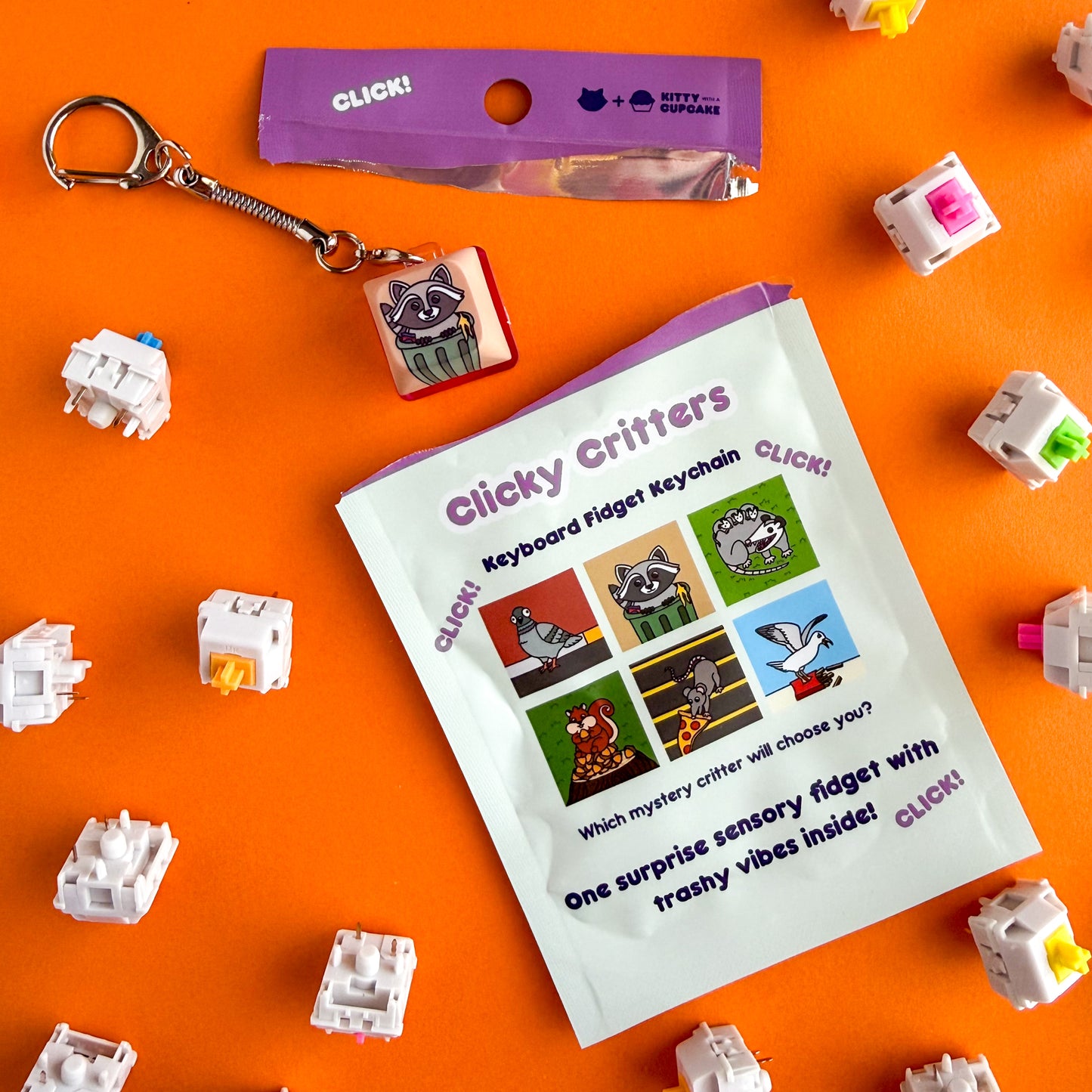 An open blind bag package with a keychain that has just been pulled from it. The bag reads "Clicky Critters - Keyboard Fidget Keychain". This is all on an orange background with keyboard switches around it. 