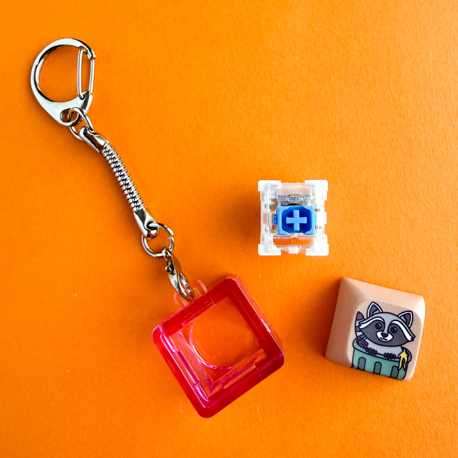 All of the pieces of the Clicky Critter Fidget Keychain, the actual keychain with the housing, the mechanical switch, and the keycap. The keycap features a cute raccoon in a trash can. 
