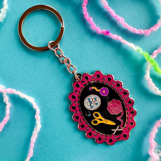 A rose gold colored metal keychain with a charm featuring an illustration that has all the tools you need work on a lace knitting project.